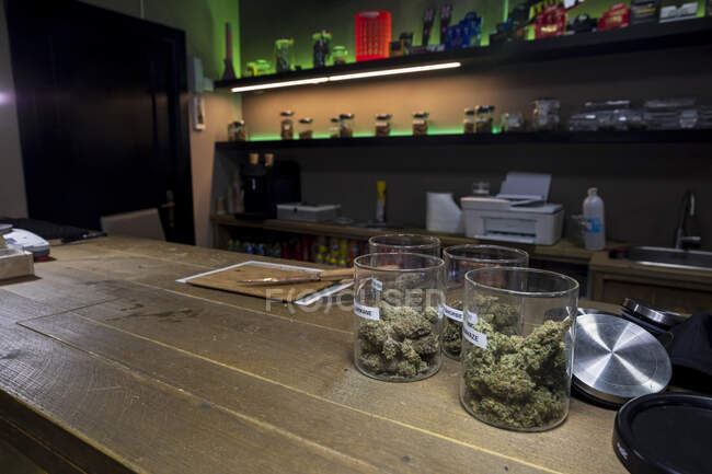 Containers of dry cannabis floral buds on table with knife and cutting board against shelves in room — Stock Photo