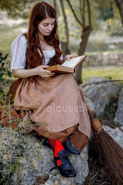Focada bruxa fêmea leitura livro mágico enquanto sentado na rocha perto de vassoura em bosques — Fotografia de Stock