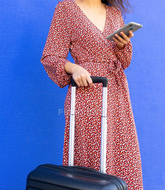 Recadré femelle méconnaissable en robe rouge longue debout avec des bagages tandis que le message de tet sur smartphone dans la rue contre le mur bleu en journée — Photo de stock