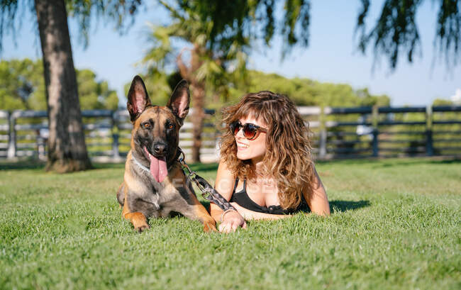 Ground level of cheerful young female in sunglasses against Malinois with tongue out lying on lawn on sunny day — Stock Photo