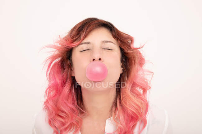 Mulher alegre com cabelo rosa soprando chiclete e olhando em frente com os olhos fechados contra fundo claro — Fotografia de Stock
