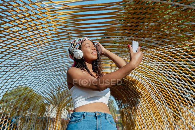 Jeune femme ethnique positive avec un téléphone portable prenant un selfie tout en écoutant la musique des écouteurs sans fil tout en riant en ville — Photo de stock