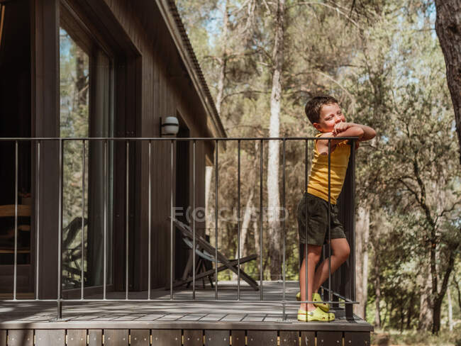 Vue latérale du petit garçon debout sur la véranda du chalet moderne situé dans les bois en été — Photo de stock