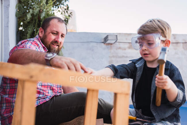 Side view of hipster dad helping son on protective glasses with hammer while doing woodwork — Stock Photo
