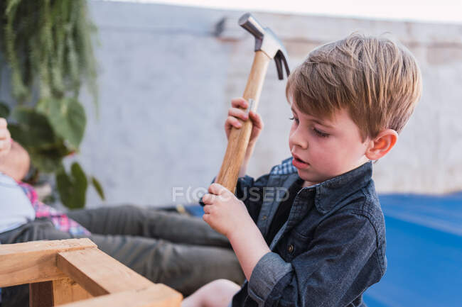 Enfant concentré en chemise denim assis avec marteau contre pièce en bois à la lumière du jour — Photo de stock