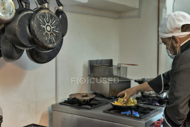 Vista lateral da cultura cozinheiro masculino étnico em máscara uniforme e estéril preparando macarrão na frigideira na cozinha do restaurante — Fotografia de Stock