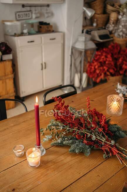 Зверху святковий стильний декоративний різдвяний букет з гілками евкаліпта і яскраво-червоними гілками з ягодами, розміщеними на дерев'яному столі зі свічками в кімнаті — стокове фото