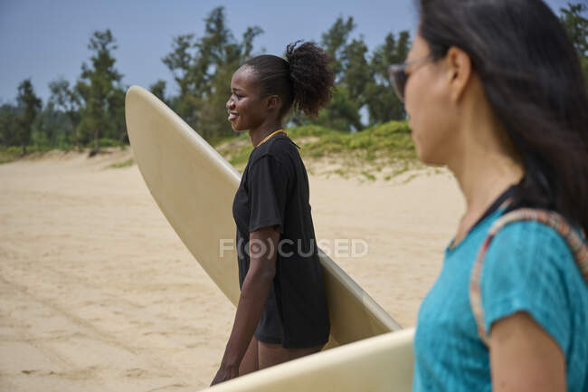 Улыбающаяся чернокожая женщина с лонгбордом против азиатской подруги с доской для серфинга, смотрящей вперед в океане под облачным голубым небом — стоковое фото