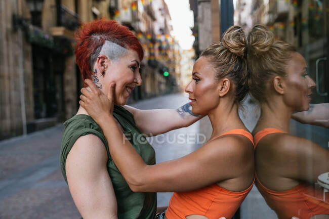 Vista lateral de la joven pareja lesbiana alegre de moda con tatuaje abrazándose mirándose en el momento del beso apoyado en una pared en la ciudad - foto de stock