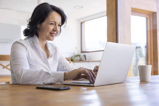 Счастливая зрелая женщина-фрилансер, просматривающая в Интернете нетбук, работающая над новым проектом, сидя дома за столом — стоковое фото