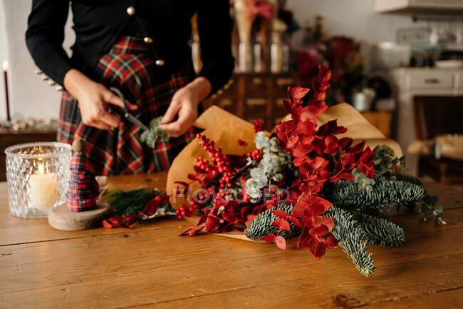 Crop unrecognizable female florist standing and cutting twigs of fir while arranging Christmas bouquet on wooden table — Stock Photo