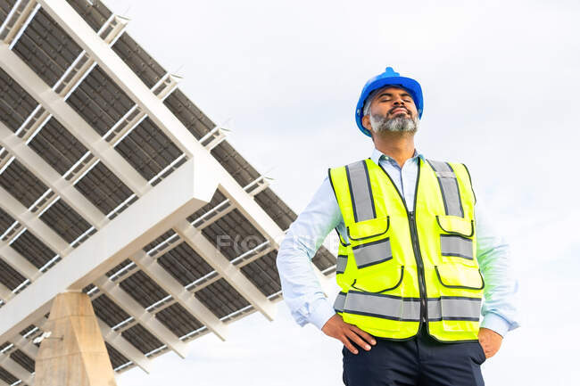 From below of mature ethnic male contractor in hardhat and vest breathing with closed eyes while standing near solar power station — Stock Photo