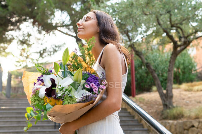 Da sotto contenuto giovane femmina in occhiali con gli occhi chiusi in piedi con mazzo di fiori in fiore sulle scale urbane — Foto stock
