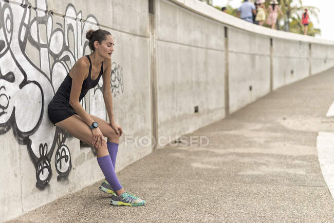 Vista lateral de la corredora atlética positiva en ropa deportiva apoyada en la pared mientras toma un descanso durante el entrenamiento en la ciudad - foto de stock