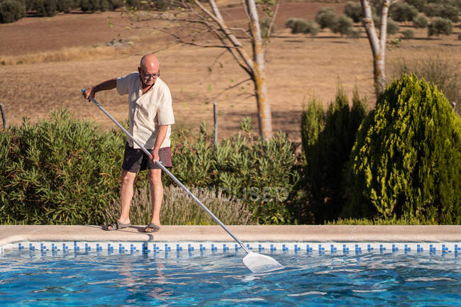 Повне тіло літнього чоловіка в окулярах очищає воду в басейні з мопедом на задньому дворі — стокове фото