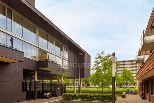 Pavimento con árboles y arbustos entre la vivienda contemporánea exterior del edificio que refleja el cielo nublado en Amsterdam Países Bajos - foto de stock