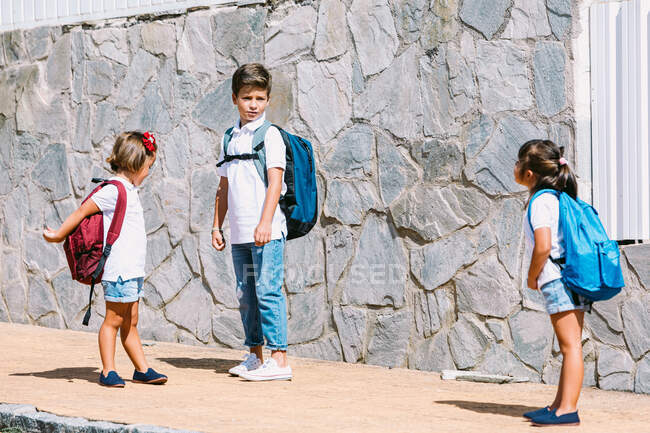 Школярка з рюкзаком розмовляє з друзями-жінками, стоячи на кахлі на кам'яній стіні на сонячному світлі — стокове фото