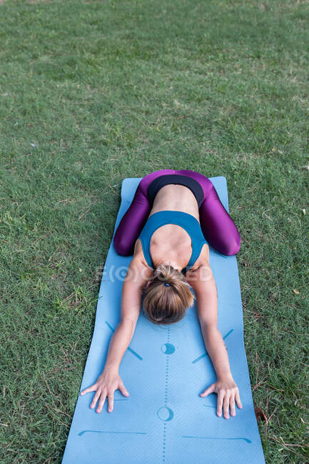 Сверху неузнаваемая молодая женщина в спортивной одежде, растягивающаяся во время занятий йогой на коврике на зеленой траве в парке при дневном свете — стоковое фото