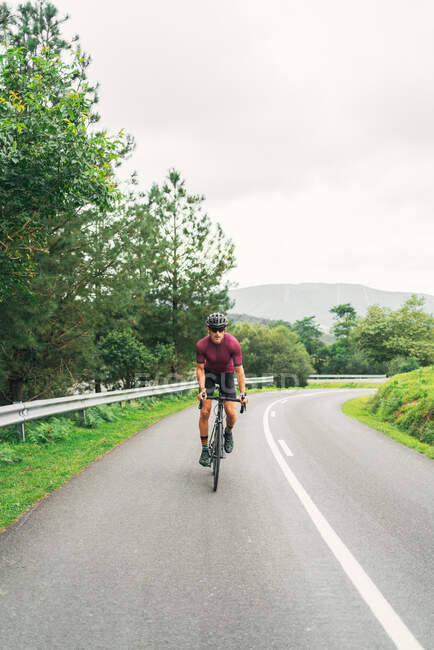 Sportsman in protective helmet riding bicycle during workout on asphalt roadway against green hill and trees under light sky — Stock Photo
