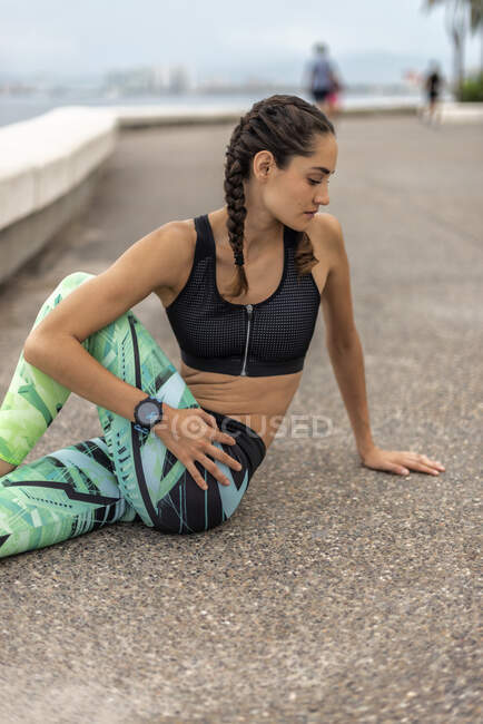 Вид сбоку на гибкую спортсменку, сидящую на набережной и растягивающую тело во время активных тренировок — стоковое фото