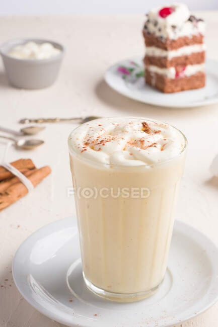 Bicchiere di latte pugno con cannella in polvere su bianco d'uovo montata contro pezzo di torta sul tavolo della mensa su sfondo chiaro — Foto stock