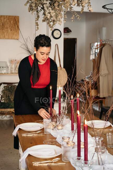 Ruhige junge Frau in legerer Kleidung steht und stellt tagsüber brennende Kerzen auf festlichen Tisch im hellen Raum — Stockfoto