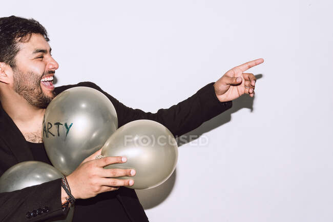 Fröhlicher bärtiger Mann mit Luftballons, der mit geöffnetem Mund lacht und während der Party vor weißem Hintergrund wegzeigt — Stockfoto