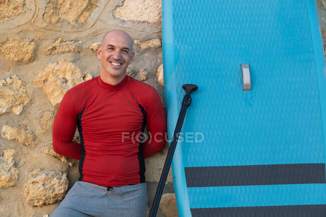 Счастливый серфер в гидрокостюме и шляпе, стоящий и смотрящий на камеру с веслом и доской SUP во время подготовки к серфингу на берегу моря, опираясь на каменную стену — стоковое фото