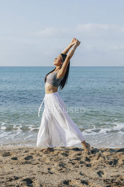 Vue latérale de la femelle ethnique penchée en arrière tout en se tenant à Ashta Chandrasana pose pendant la pratique du yoga sur la côte sablonneuse contre l'océan — Photo de stock
