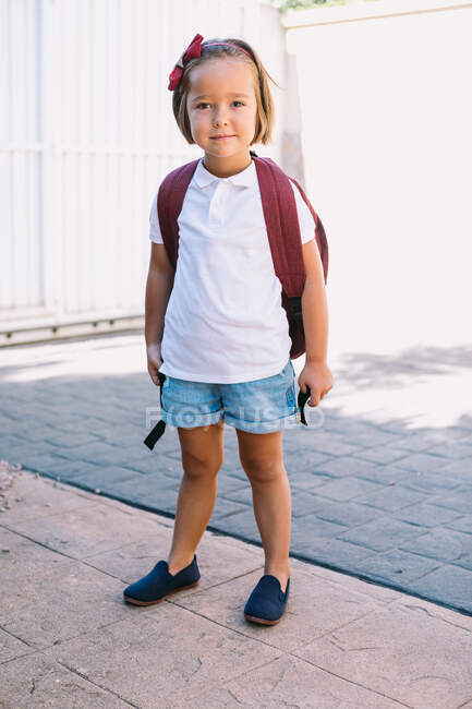 Schulkind mit Rucksack steht auf Gehweg und blickt im Sonnenlicht in Kamera — Stockfoto