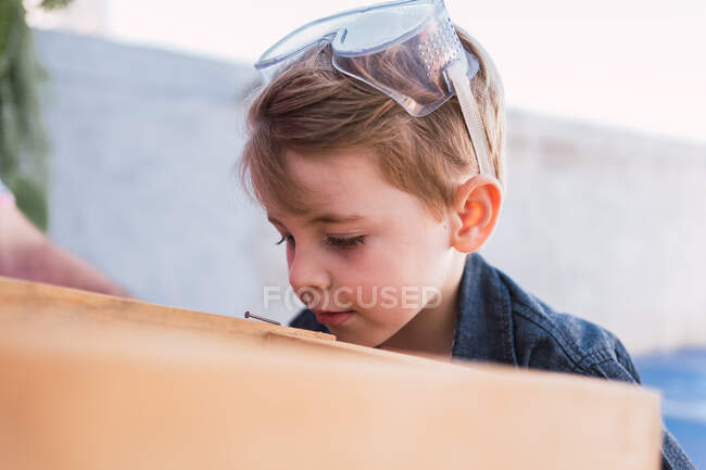 Contemplar al niño con gafas de seguridad y camisa de mezclilla mirando hacia otro lado contra el taburete hecho a mano durante el día - foto de stock