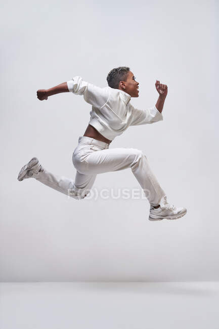 Vista lateral da jovem fêmea afro-americana rápida em roupas da moda correndo e pulando acima da superfície clara — Fotografia de Stock