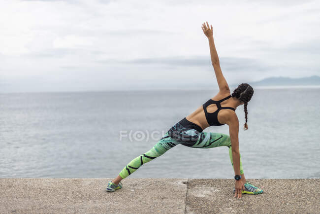 Vue de dos d'une sportive flexible faisant de l'exercice de flexion et réchauffant son corps pendant l'entraînement sur la promenade sur fond de mer — Photo de stock