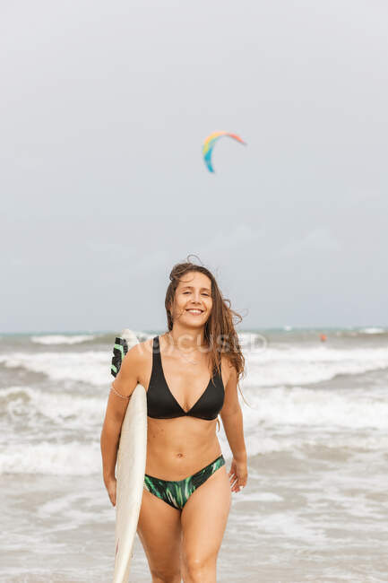 Jovem desportista alegre com cabelo voador e prancha de surf no oceano com espuma sob céu nublado — Fotografia de Stock