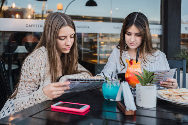 Best female friends reading menu at cafeteria table with cocktails and pastries on plate against smartphone in town — Stock Photo