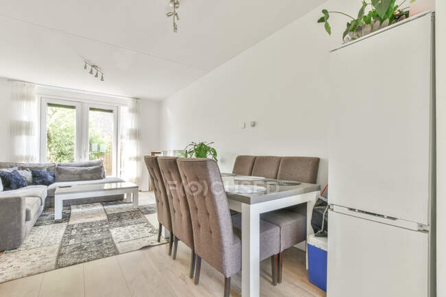 Innenraum eines hellen Zimmers mit Esstisch und Stühlen gegen graues Sofa mit Kissen und Teppich tagsüber — Stockfoto