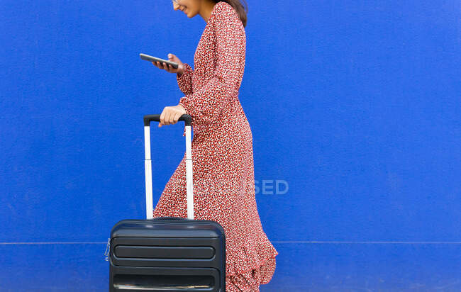 Side view of cropped unrecognizable female in long red dress walking with luggage while tet message on smartphone on the street against blue wall in daytime — Stock Photo