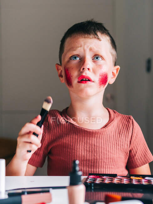 Charming child with makeup applicator looking up while sitting at table with eyeshadow palette — Stock Photo