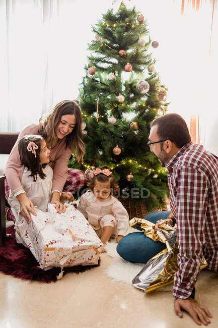 Los padres sonrientes con sus hijas abren cajas de regalo en el suelo mientras celebran las vacaciones de Año Nuevo en casa - foto de stock