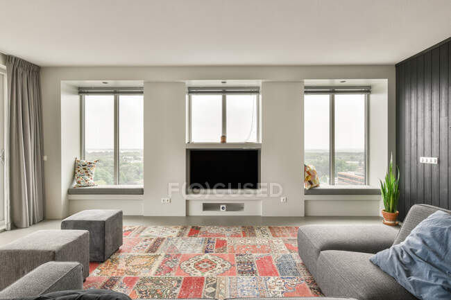 Современная гостиная с телевизором и окнами, расположенными напротив мягкого дивана и цветного ковра в квартире днем — стоковое фото