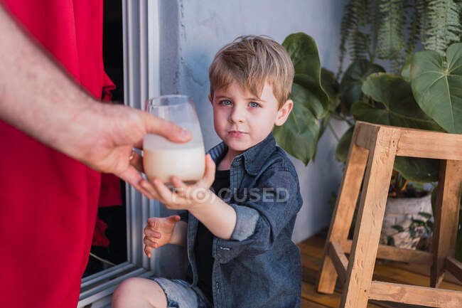 Child looking at camera while receiving glass of beverage from crop unrecognizable dad against handmade stool — Stock Photo
