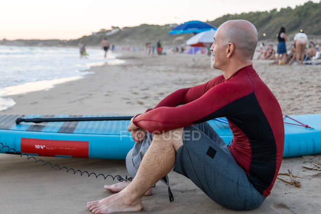 Side view of thoughtful male surfer in wetsuit sitting looking away with SUP board while preparing to surf on seashore — Stock Photo