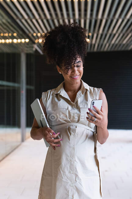 Приятная беременная афроамериканка с кудрявыми волосами и текстовыми сообщениями на мобильном телефоне во время ношения ноутбука возле стеклянного здания — стоковое фото