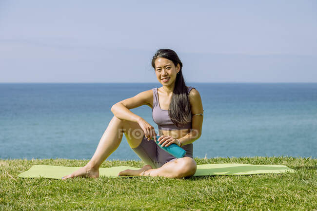 Allegro giovane femmina etnica a piedi nudi in abbigliamento sportivo con bottiglia d'acqua seduta contro l'oceano e guardando la fotocamera — Foto stock