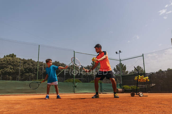 Adult man showing boy how to hold paddle and hit ball during tennis training on sunny day on court — Stock Photo