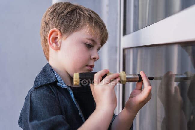 Side view of focused child learning how to use screwdriver while reflecting in window in daytime — Stock Photo