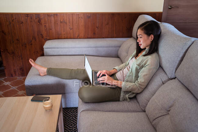 Вид сбоку заинтересованной молодой сотрудницы, работающей над нетбуком на диване против кофе и смартфона дома — стоковое фото