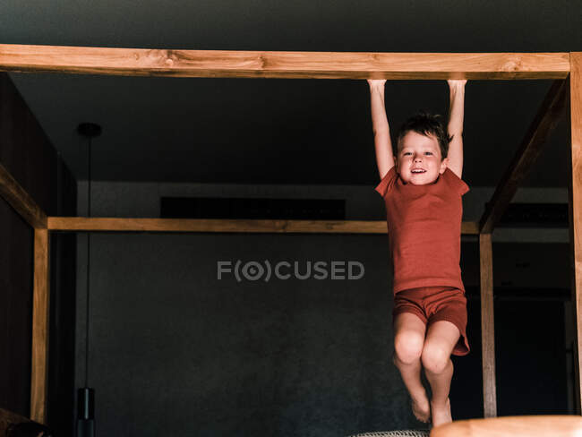 Веселый маленький ребенок, висящий на деревянной кровати с балдахином, развлекаясь в спальне и глядя в камеру — стоковое фото