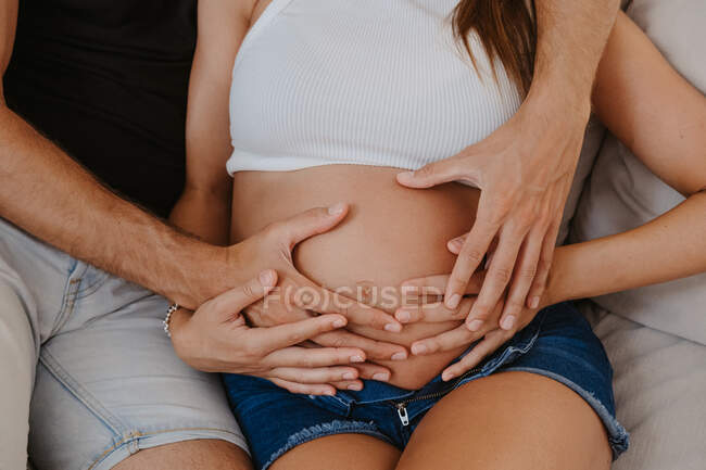 Cultivé homme méconnaissable embrassant ventre de femme enceinte bien-aimée tout en se reposant sur le canapé dans le salon — Photo de stock