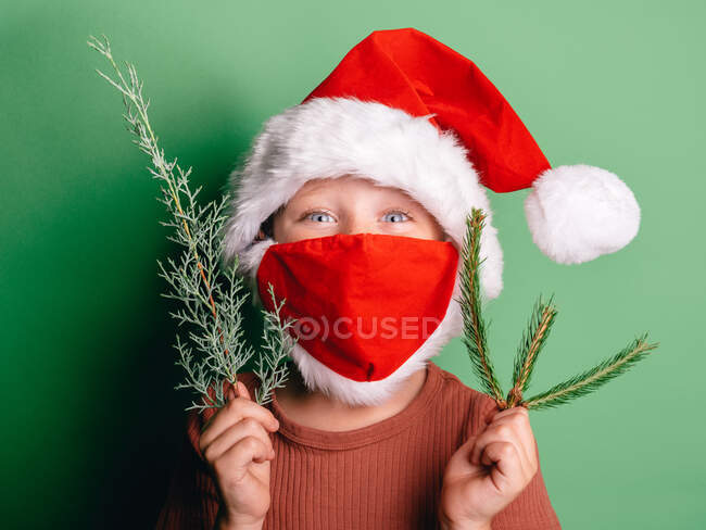 Boy in red Santa hat and mask for protecting from COVID with fir branches looking at camera with eyes wide open — Stock Photo